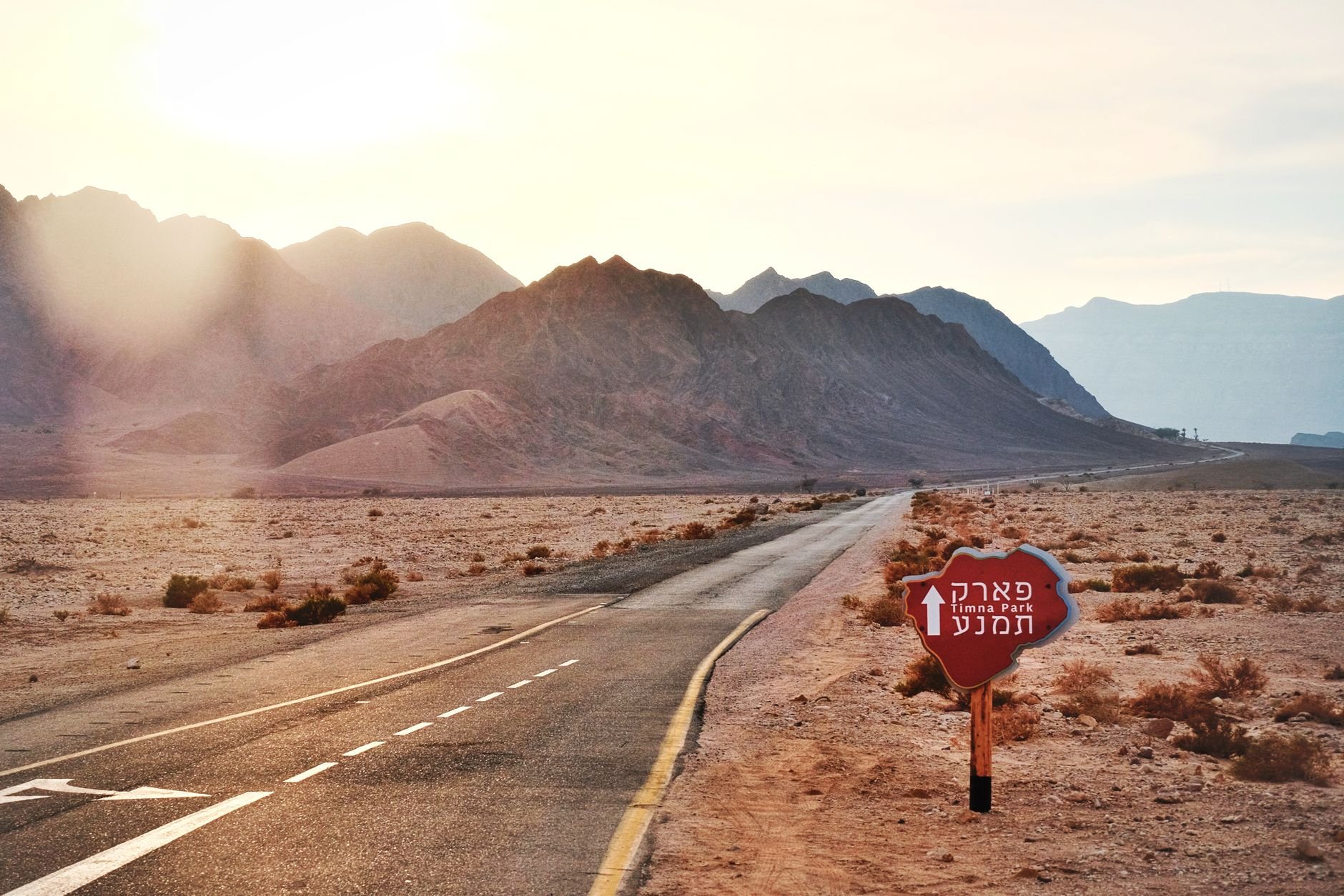 Empty road leading to Timna Park with rugged mountains and desert terrain under warm sunlight.